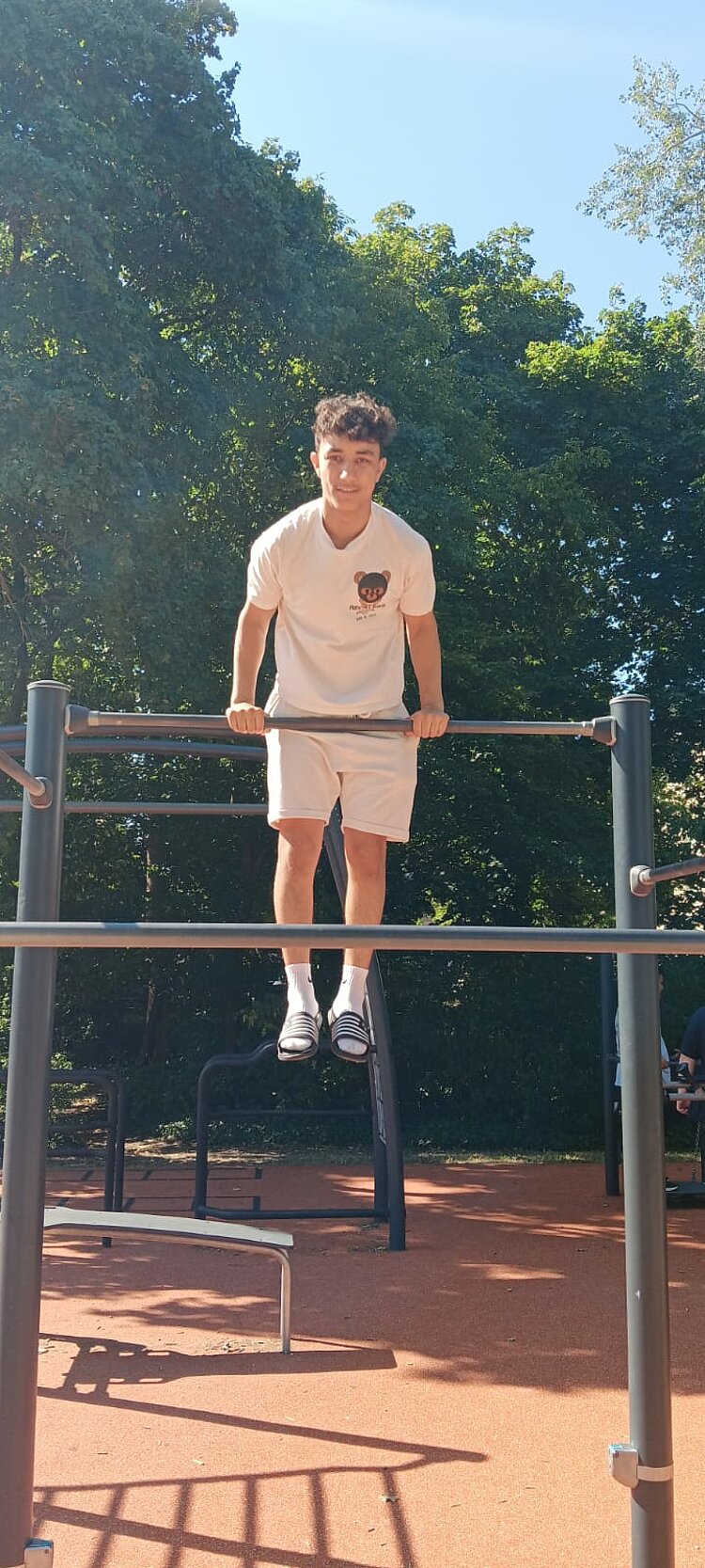 Ein junger Mann trainiert an einer Outdoor-Fitnessanlage und stützt sich mit gestreckten Armen auf einer hohen Metallstange ab.