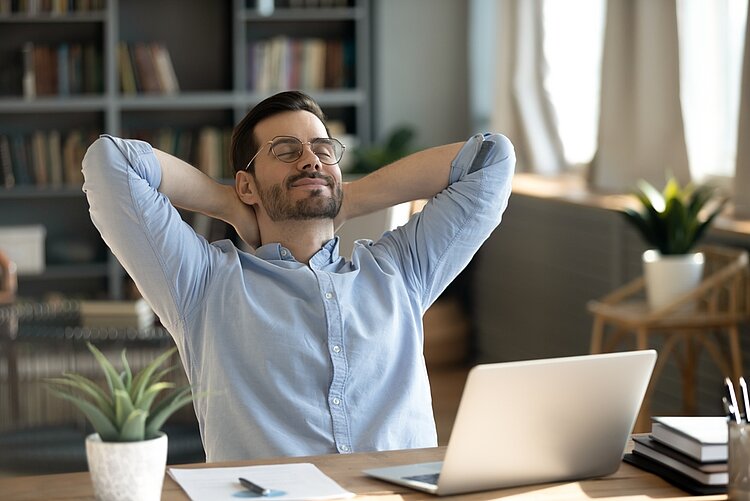 An einem Schreibtisch mit Laptop sitzt ein Mann in hellblauem Hemd. Der Mann lehnt sich mit geschlossenen Augen und hinter dem Kopf verschränkten Armen zurück und lächelt dabei.