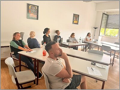 Teilnehmende sitzen in einem Schulungsraum an Tischen und verfolgen aufmerksam eine digitale Präsentation.
