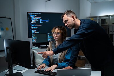 Ein Mann unterstützt eine junge Frau an einem Computerarbeitsplatz und zeigt auf den Bildschirm. Im Hintergrund sind mehrere Monitore mit Programmcode zu sehen.