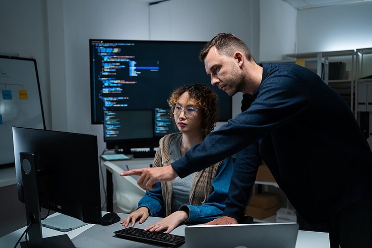 Ein Mann unterstützt eine junge Frau an einem Computerarbeitsplatz und zeigt auf den Bildschirm. Im Hintergrund sind mehrere Monitore mit Programmcode zu sehen.