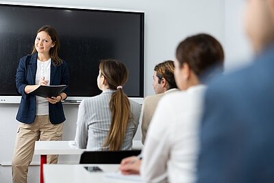 Eine Frau steht vor einer Gruppe von Menschen in einem modernen Seminarraum. Sie trägt einen dunkelblauen Blazer und hält einen Ordner in den Händen. Im Hintergrund befindet sich ein großer, ausgeschalteter Flachbildschirm an der Wand. Die Teilnehmenden sitzen an weißen Tischen und hören aufmerksam zu. Die Szene wirkt sachlich und konzentriert.