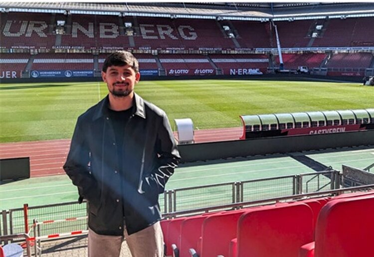 Ein männlicher Teilnehmender der Werkstattschule des Euro-Bildungswerkes Nürnberg steht lächelnd auf der Tribühne im Fußballstadion und posiert für ein Foto.