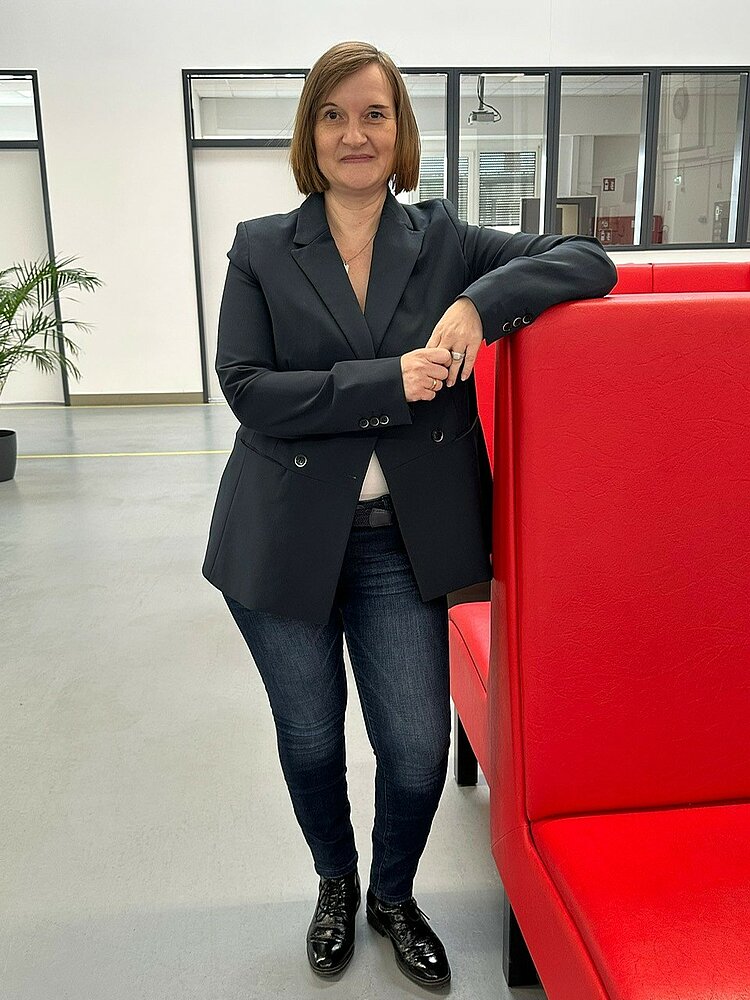 Lydia Olschock, die Schulleiterin der EBW Nürnberg, steht in einem modernen Büro oder öffentlichen Raum. Sie trägt ein dunkles Sakko, ein weißes Oberteil, Jeans und schwarze Schuhe. Sie lehnt mit einem Arm an einer roten Sitzbank und lächelt in die Kamera.