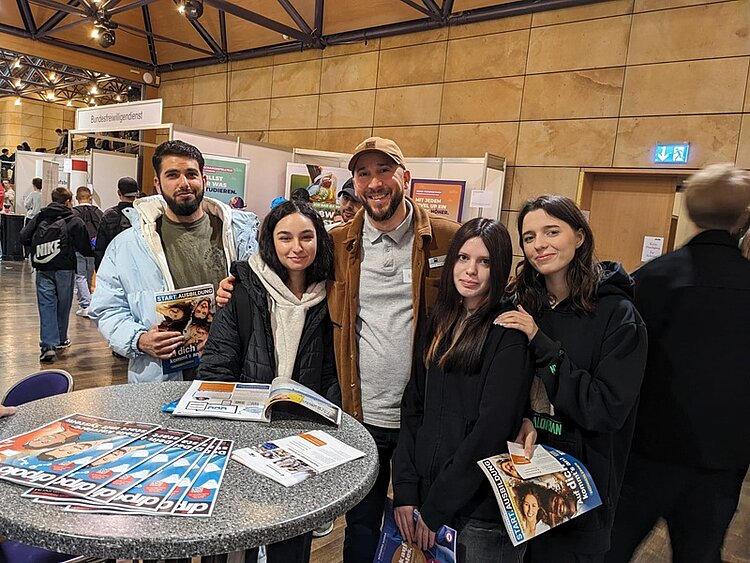Werkstattschule auf der vocatium Fürth 2025 Die Teilnehmenden der Werkstattschule des Euro-Bildungswerkes Nürnberg auf der vocatium Messe posieren an einem Stehtisch, der voller Informationsbroschüren ist, für ein Gruppenfoto.
