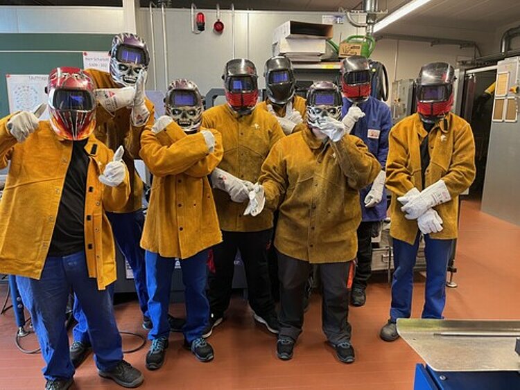Eine Personengruppe mit Arbeitsschutzkleidung für Schweißarbeiten und Schutzhelmen mit heruntergeklappten Visier, auf denen zum Teil eine Zeichnung von einem Totenkopf zu sehen ist, steht in einem Werkstattraum.