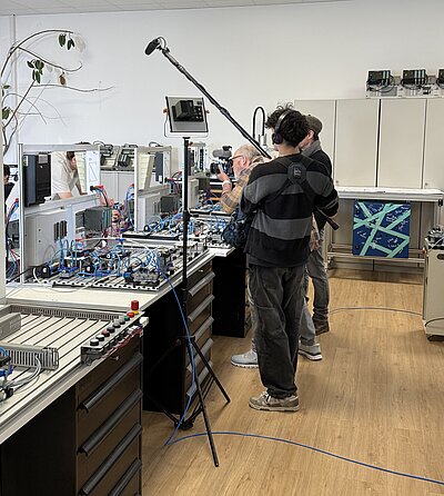 Drehtag in einer Werkstatt mit Kamera und Mikrofon. Mehrere Personen filmen an industriellen Maschinen und Technikaufbauten.