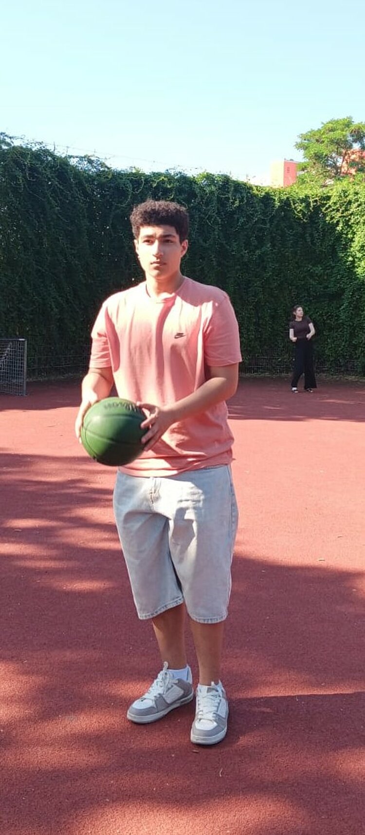 Ein junger Mann steht auf einem Sportplatz und hält einen grünen Basketball vor sich; im Hintergrund ist eine weitere Person zu sehen.