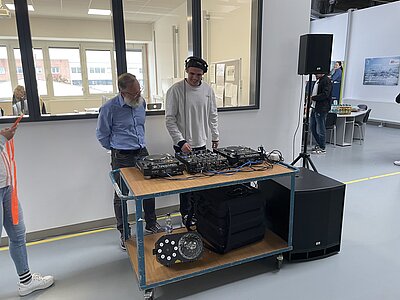 Halloween-Feier Ein DJ-Pult in einer Halle. Dahinter stehen zwei Männer. Der rechte bedient das Pult und der linke Mann guckt interessiert zu.