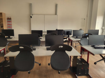 Heller Computerraum mit mehreren Arbeitsplätzen, schwarzen Monitoren und Bürostühlen. An der Stirnseite des Raums befindet sich ein Whiteboard.