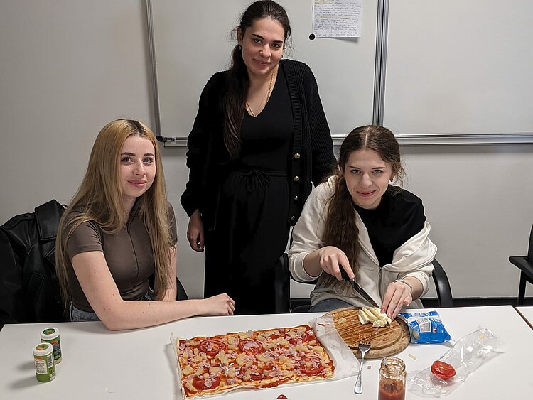 Drei Frauen sitzen und stehen um einen Tisch und schneiden eine Pizza.