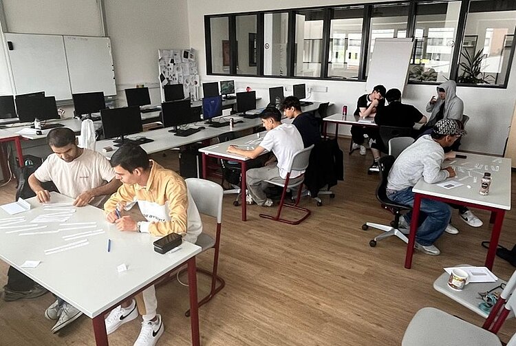 Mehrere junge Menschen sitzen an einzelnen Tischen in einem modernen Klassenraum. Sie arbeiten konzentriert mit Papierstreifen oder Arbeitsblättern. Im Hintergrund stehen Computer auf Tischen entlang der Wand. Die Atmosphäre wirkt ruhig und fokussiert. Einige Personen unterhalten sich in kleinen Gruppen, während andere allein arbeiten. Die Einrichtung ist schlicht, mit weißen Wänden, großen Fenstern und Holzboden.