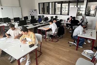 Mehrere junge Menschen sitzen an einzelnen Tischen in einem modernen Klassenraum. Sie arbeiten konzentriert mit Papierstreifen oder Arbeitsblättern. Im Hintergrund stehen Computer auf Tischen entlang der Wand. Die Atmosphäre wirkt ruhig und fokussiert. Einige Personen unterhalten sich in kleinen Gruppen, während andere allein arbeiten. Die Einrichtung ist schlicht, mit weißen Wänden, großen Fenstern und Holzboden.