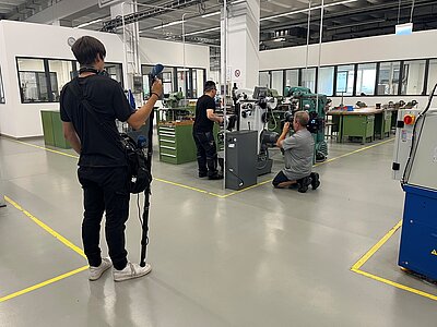 Fernsehdreh zum Thema Bürgergeld am Standort des EBW Nürnberg Eine Lernwerkstatt mit mehreren Arbeitsplätzen. Ein Mann bedient eine der Maschinen während er von einem daneben hockenden Mann mit einer Fernsehkamera gefilmt wird. Links davon steht ein schwarz gekleideter Mann mit Mikrofon und weiterem Equipment für die Aufzeichnung