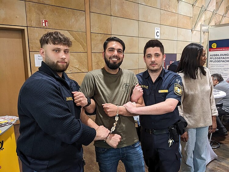 Werkstattschule auf der vocatium Fürth 2025 Ein Teilnehmer der Werkstattschule des Euro-Bildungswerkes Nürnberg auf der vocatium Messe lässt sich am Stand der Polizei anhand seiner selbst vorführen, wie eine Verhaftung, inklusive Handschellen, vonstatten geht. Der Teilnehmer steht zwischen beiden jungen Polizisten, alle posieren für ein gemeinsames Foto.
