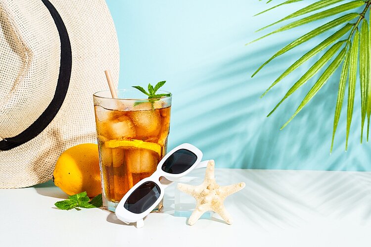 Vor hellblauen Hintergrund liegt ein Strohhut, eine Sonnenbrille und ein mit Eis gefülltes Glas.