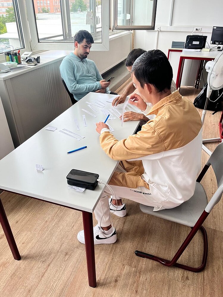 Im Vordergrund sitzen zwei Personen an einem rechteckigen Tisch und sortieren weiße Papierstreifen. Beide tragen helle Kleidung mit beigen und weißen Farbakzenten. Auf dem Tisch liegen mehrere Stifte, ein schwarzes Etui und weitere Papierstreifen. Im Hintergrund sitzt eine weitere Person an einem separaten Tisch und blickt auf ein Smartphone. Der Raum ist hell, mit großen geöffneten Fenstern, weißen Wänden und einem Holzfußboden. Ein Ventilator steht neben dem Tisch.