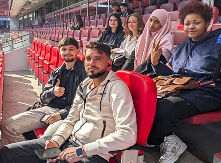 Die weinlichen und männlichen Teilnehmenden der Werkstattschule des Euro-Bildungswerkes Nürnberg sitzen lächelnd auf den roten Plastikstühlen im Fussballstadion und posiert für ein Foto.