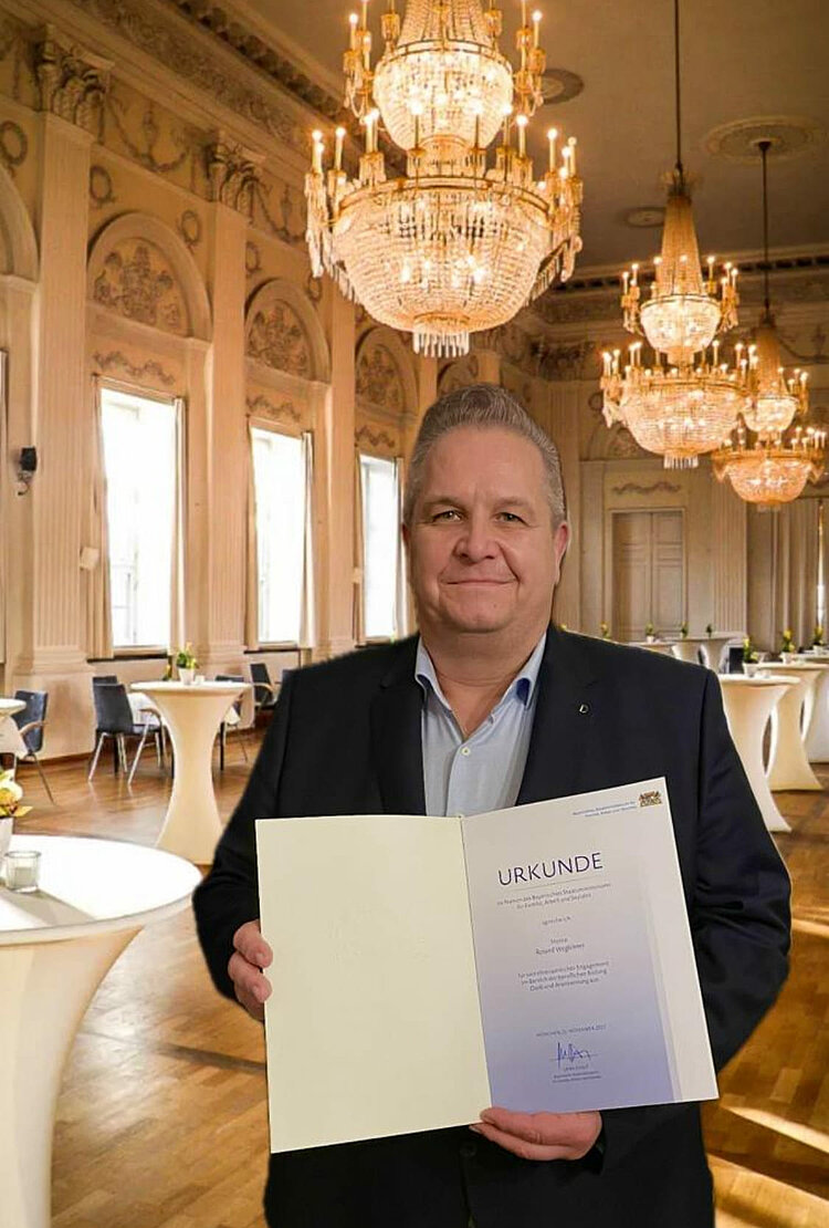 Auszeichnung für Ehrenamtliche Ein Mann blickt freundlich in die Kamera, in seinen Händen hält er eine Urkunde zu seiner ehrenamtlichen Tätigkeit. Im Hintergrund ist ein prunkvoller, goldener Saal mit Kronleuchtern und Stehtischen zu sehen.