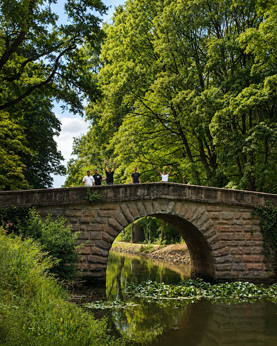 Fünf Personen stehen auf einer steinernen Bogenbrücke in einem grünen Park. Zwei von ihnen heben die Arme, unter der Brücke fließt ein schmaler Wasserlauf.