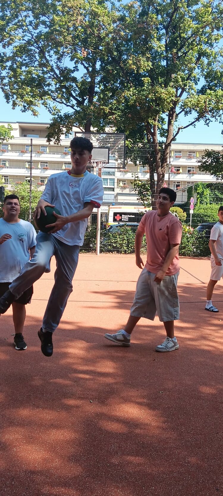 Mehrere junge Männer spielen auf einem Outdoor-Sportplatz mit einem grünen Ball; ein Spieler hält den Ball während einer dynamischen Bewegung in den Händen.