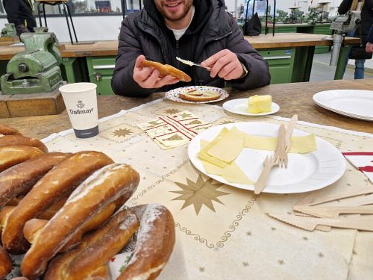 Weihnachtsfeier der Umschüler Ein Mann sitzt an einem Tisch und bestreicht eine Brezel mit Butter. Im Vordergrund sind mehrere Brezeln zu sehen und einen Teller mit Käsescheiben und Holzgabeln. Auf dem Tisch stehen außerdem ein Pappbecher und ein Teller mit Butter. Die Szene scheint sich in einem Werkraum abzuspielen, denn im Hintergrund befinden sich Werkbänke mit Schraubstöcken.