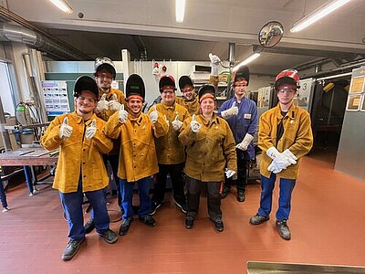 Eine Gruppe von jungen Personen mit hochgeklappten Schutzhelmen, gelben Schutzjacken und weißen Schutzhandschuhen für Schweißarbeiten stehen in einem Werkstattraum und machen das Gefällt-mir-Zeichen mit dem Daumen.