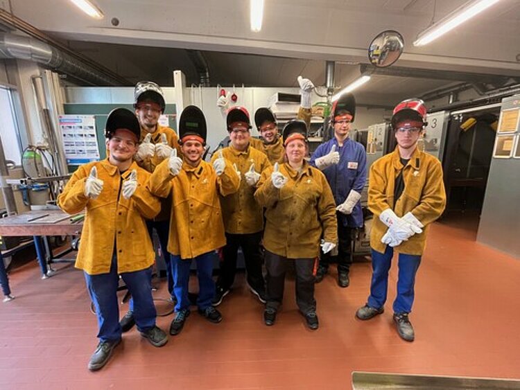 Eine Gruppe von jungen Personen mit hochgeklappten Schutzhelmen, gelben Schutzjacken und weißen Schutzhandschuhen für Schweißarbeiten stehen in einem Werkstattraum und machen das Gefällt-mir-Zeichen mit dem Daumen.